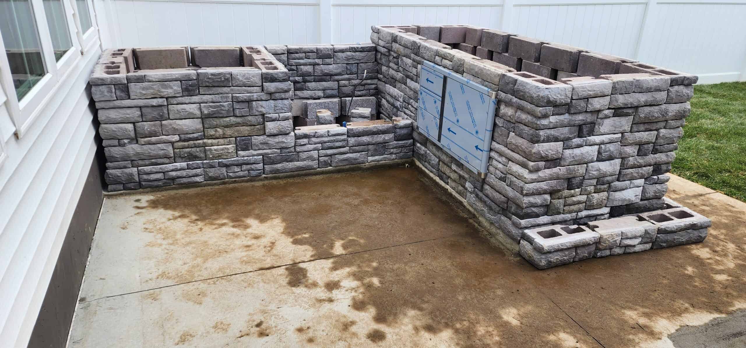 a stone outdoor kitchen and firepit under construction on a large backyard patio