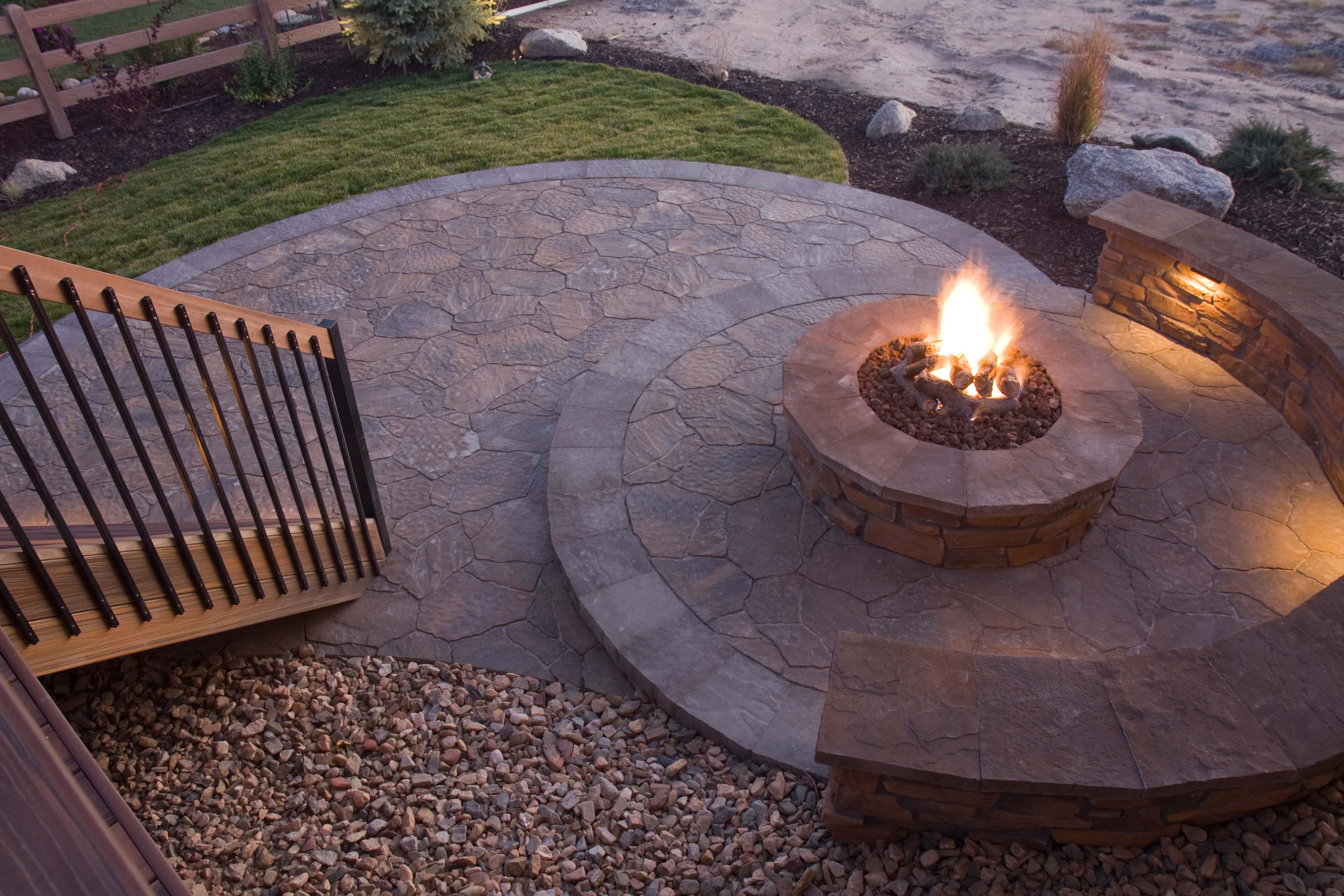 a lush mountain landscape with a rounded natural stone firepit in the center of a paver patio surrounded by landscaping